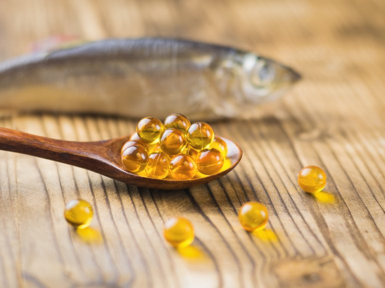 Fish oil capsules in a wooden spoon on a wooden table with a fish in the background. Selective focus. Traditional medicine.